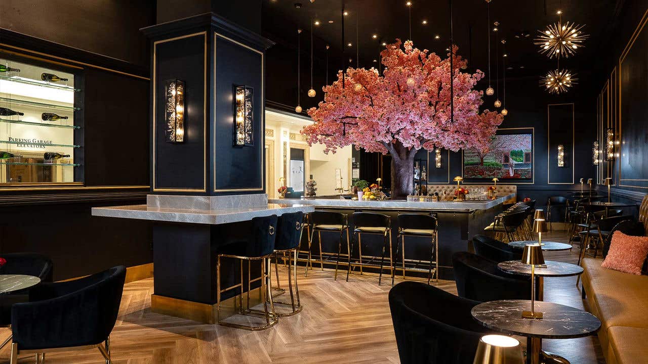 An interior of a bar with black walls and gold accents, and marble counters and bartops, with a central bar to the right with a cherry blossom tree in the middle of it