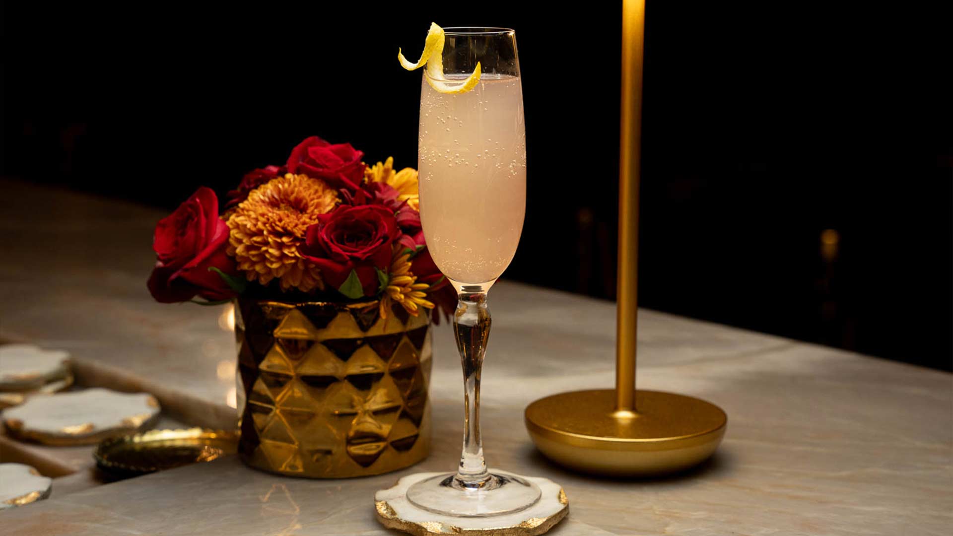A white cocktail in a champagne flute garnished with a lemon peel, on a marble bartop with a gold vase and light behind it