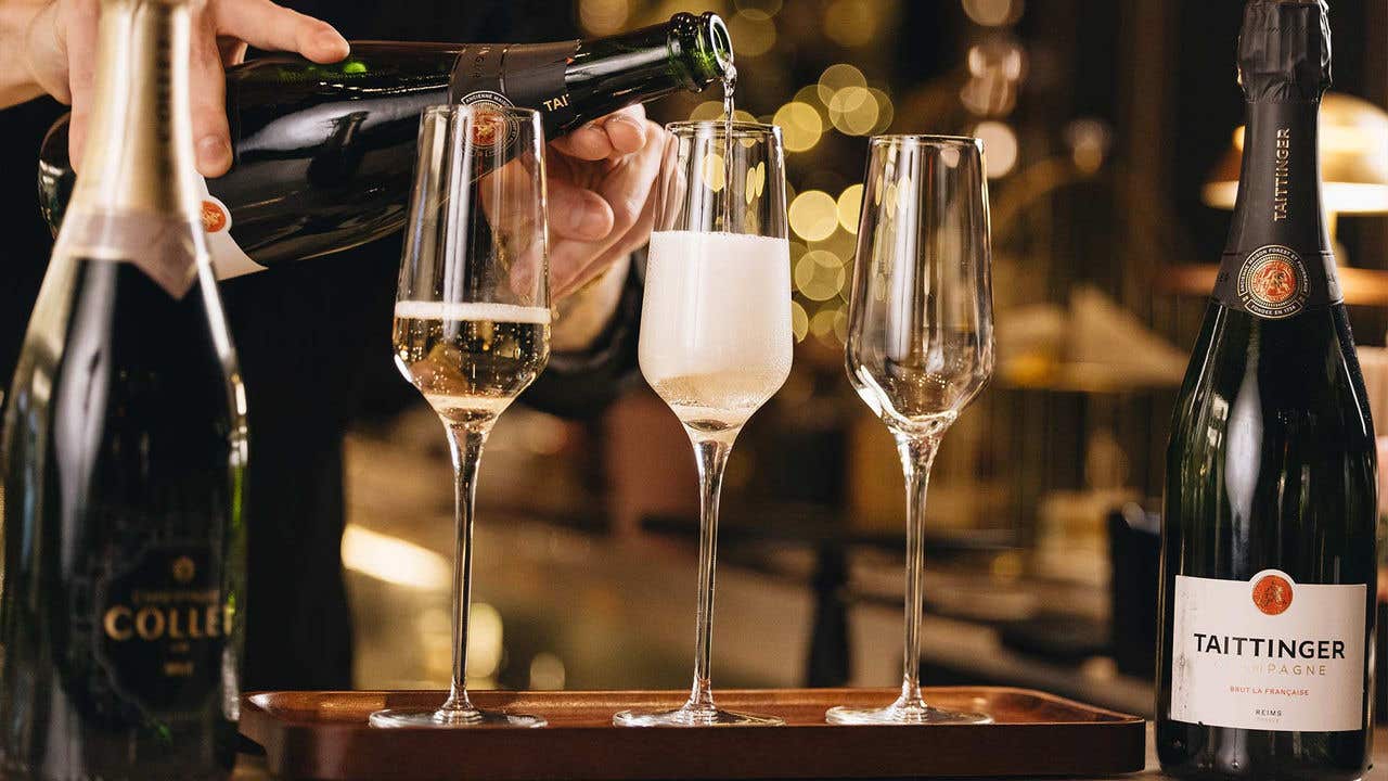A set of champagne flutes with champagne being poured into the flutes, amongst 2 bottles at the left and right