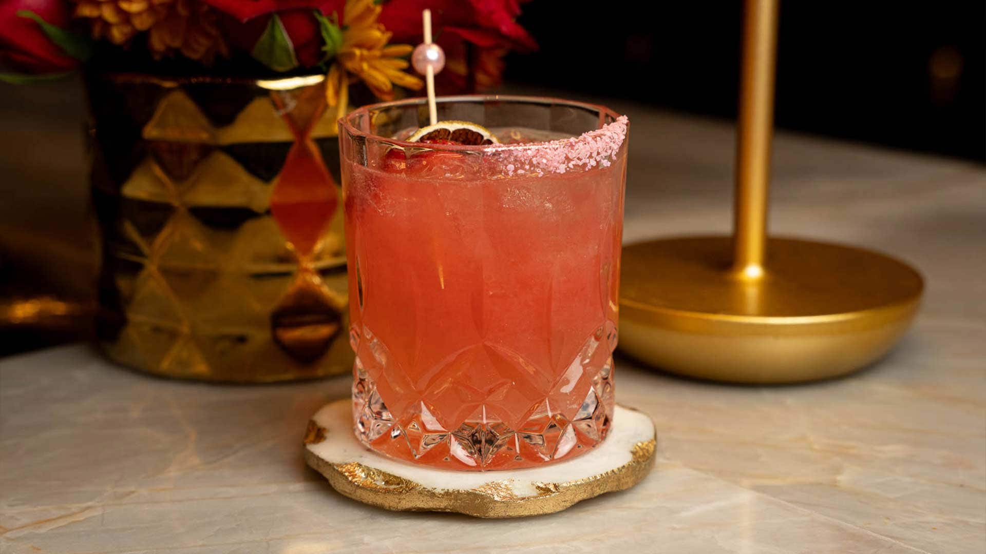 A pink cocktail in a rocks glass on a marble tabletop or bar, with a gold vase and light in the background 