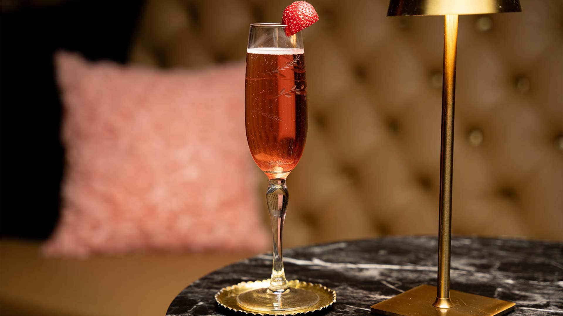 A red cocktail in a champagne flute, with bubbles at the top, and garnished with a strawberry